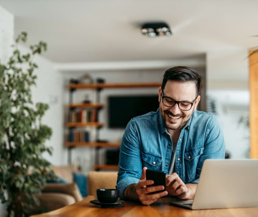 Usmievajúci sa muž s okuliarmi surfuje na smartfóne v modernom interiéri bytu.