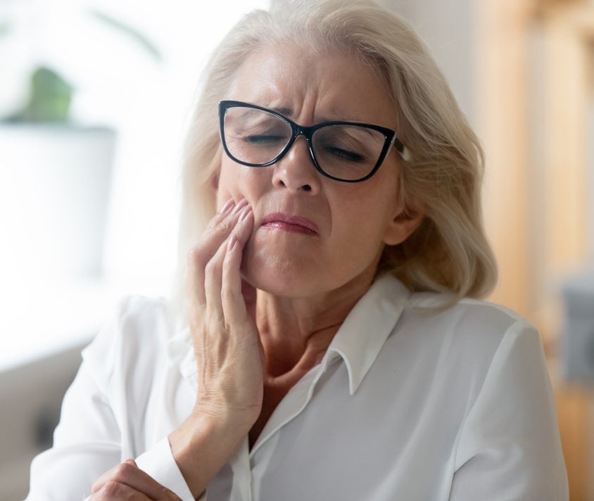Staršia žena s okuliarmi sa dotýka tváre a vyjadruje bolesť alebo nepohodlie.