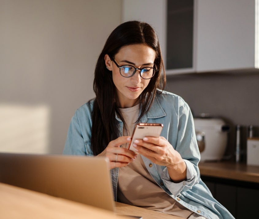 Sústredená žena v svetlej kuchyni používajúca smartfón pri stole s otvoreným notebookom.